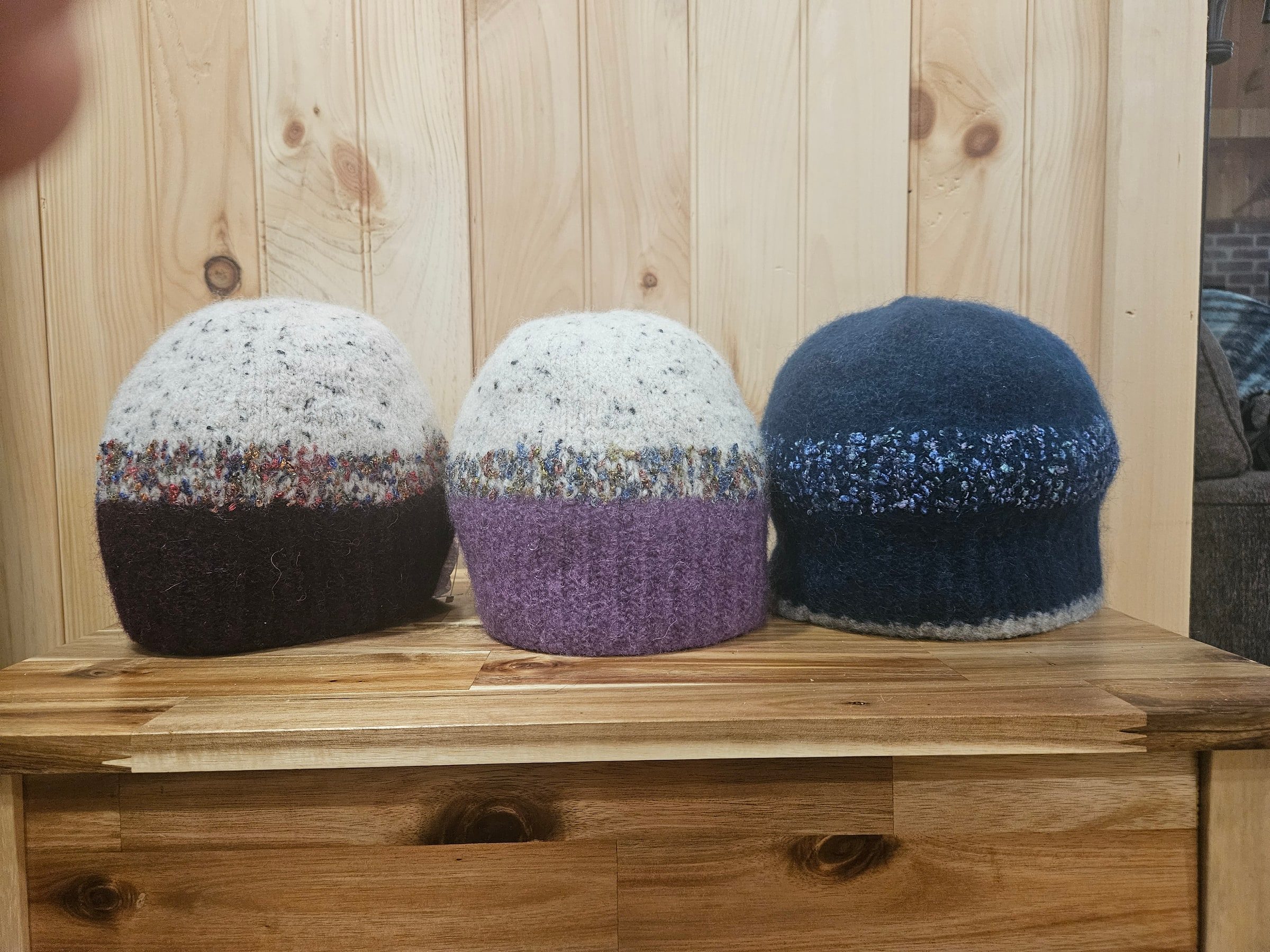 Three felted hats in burgundy, purple, and navy on a wooden bench against a pine wall.