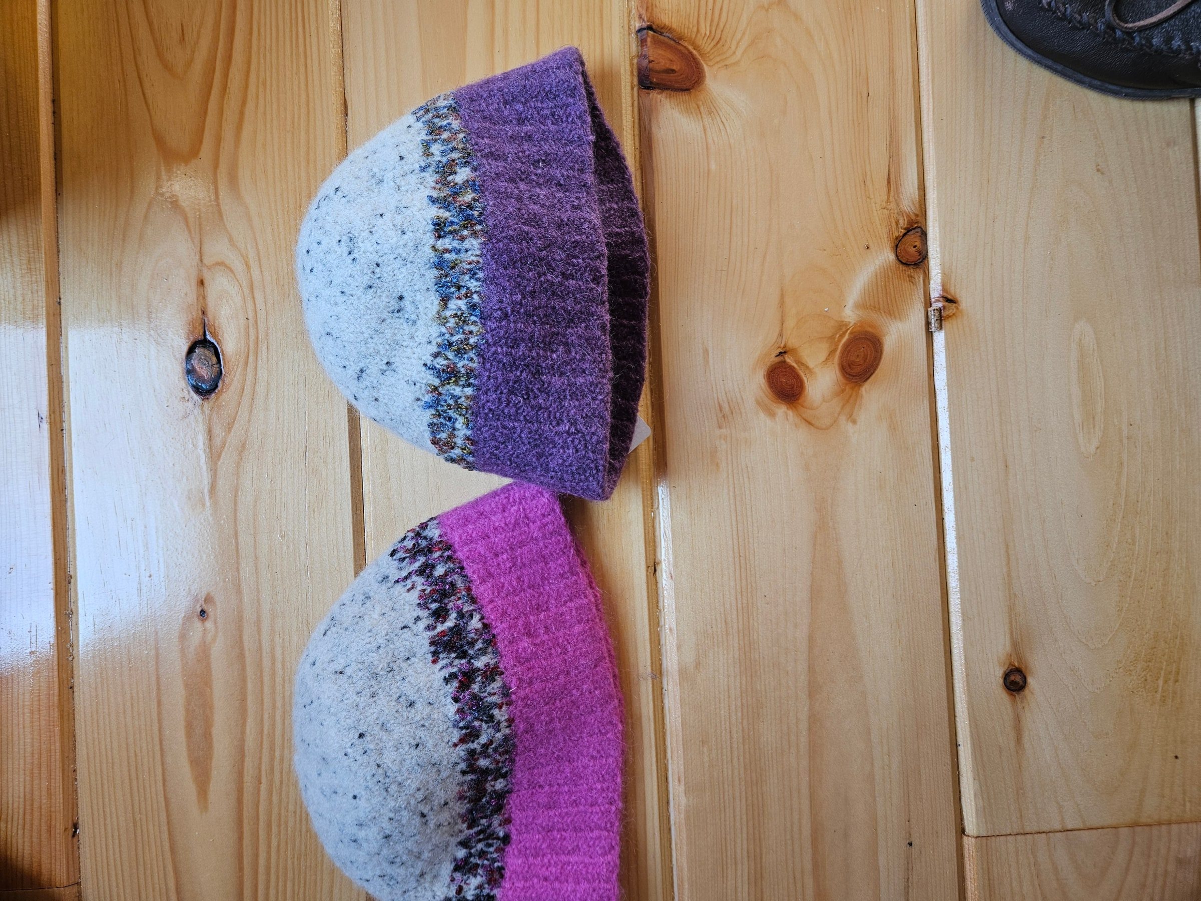 Two felted hats lying flat on a wood floor, one purple and cream, one magenta and cream.