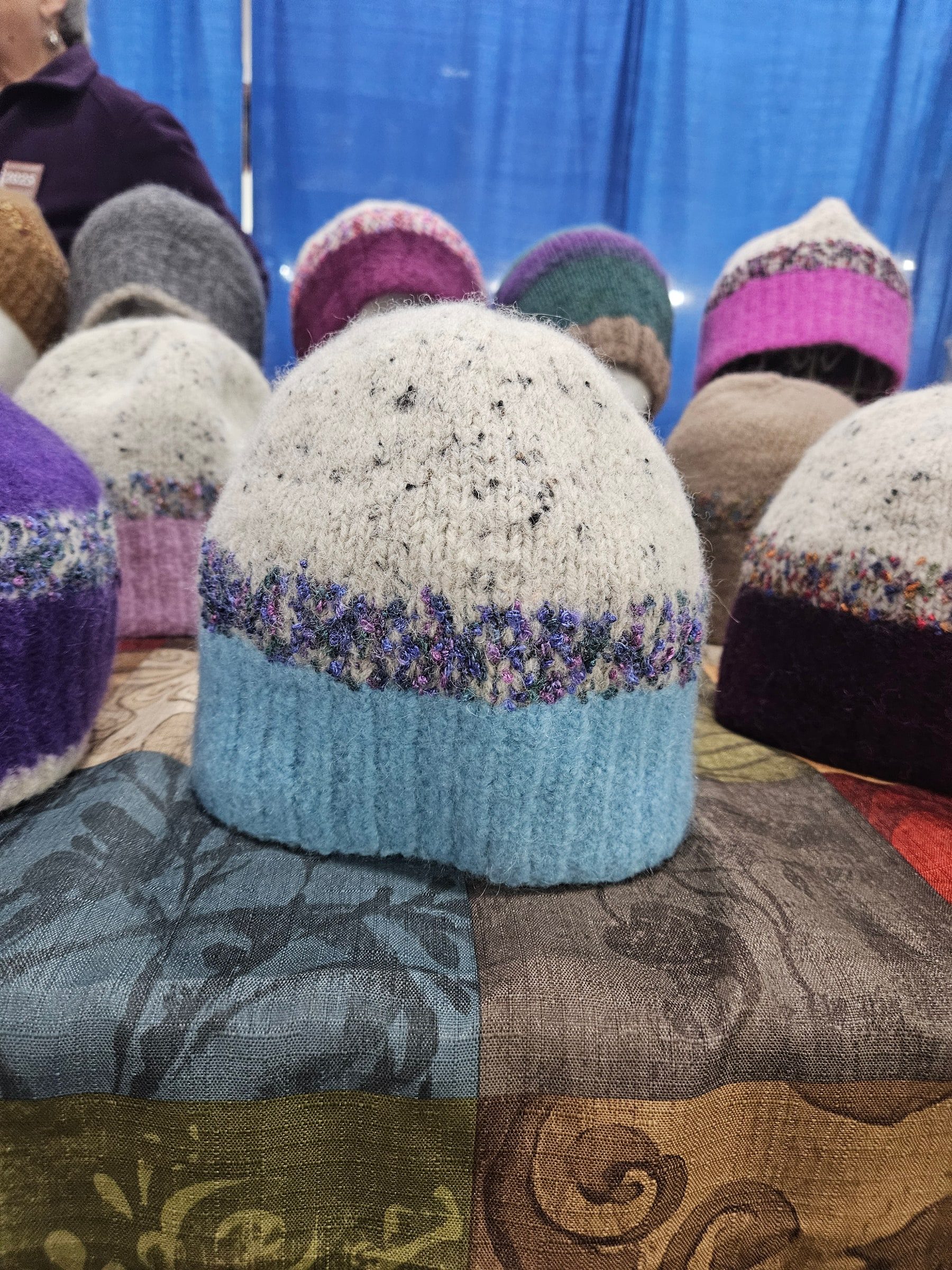 A light blue felted hat with a cream crown and multicolored band in the foreground of a craft-show display.