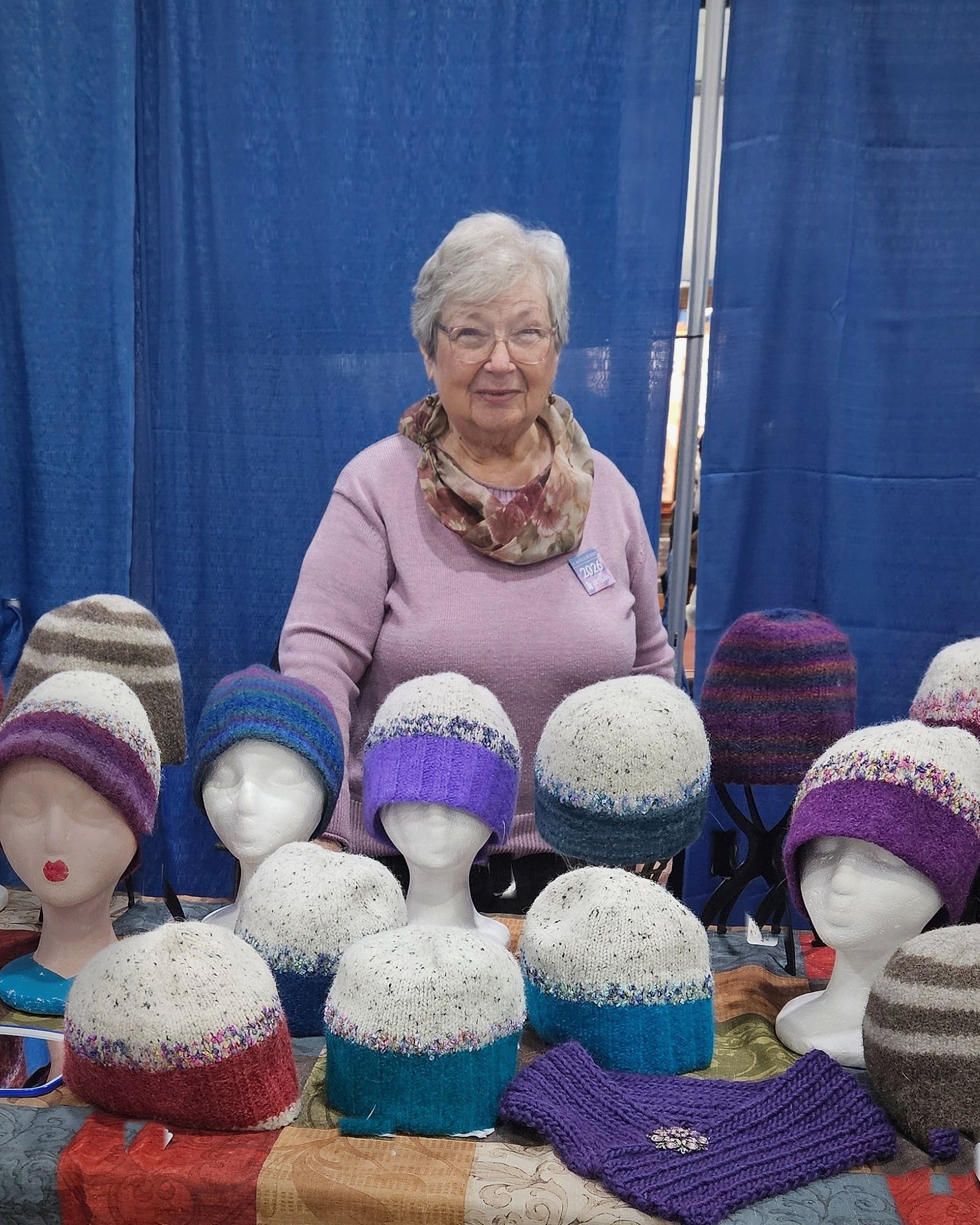 Judy Hella standing behind a table of felted wool hats at a Vermont craft show, in front of blue curtains.